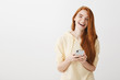 © Cookie Studio - What beautiful day to chat online. Indoor shot of emotive happy redhead woman holding smartphone, gazing at camera, laughing and being in great mood while standing over gray background