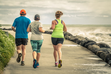 Three people running along the ocean. Two girls and one man jogging together on a beautiful road along the sea with big waves and spray. A beautiful day of outdoors sport in New Zealand.