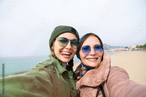 Poster Mother and adult daughter are doing selfie on the beach going crazy and showing tongueSunset light