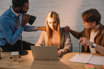 two multicultural businessmen threatening and shine lamp in face of businesswoman using laptop