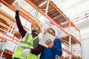 Wall Mural - Low angle portrait of warehouse manager holding clipboard talking to worker wearing hardhat and reflective jacket while discussing stock inventory, pointing up