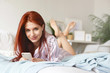 © shurkin_son - Close up of stylish young woman with ginger hair and freckled skin lying on stomach on undone bed, keeping feet in air, having playful look, smiling, flirting with some guy using app on cell phone