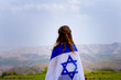 Little patriot jewish girl standing and  enjoying great view on the sky, spring field and mountains with the flag of Israel wrapped around her. Memorial day-Yom Hazikaron and Yom Ha’atzmaut concept.