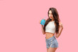Young slim brunette in denim shorts and top having refreshing drink posing flirty on pink backdrop.