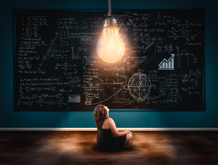 Young girl looking up to a lightbulb in front a blackboard full of formulas . The concept of solving a math formula.