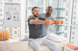 © zinkevych - Rehabilitation exercise. Nice pleasant handsome man sitting on the medical couch and holding dumbbells in front of him