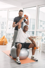 Training equipment. Nice young man sitting on the medball and lifting dumbbells