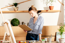 Young Female Artist Reading Book And Standing At Table With Painting Supplies