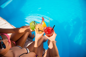 Sticker - High angle cropped shot of three girls cheering with drinks near swim pool. Red, green and yellow different cold cocktails with ice cubes, black straws, transparent clean blue aqua in pool, sun shines