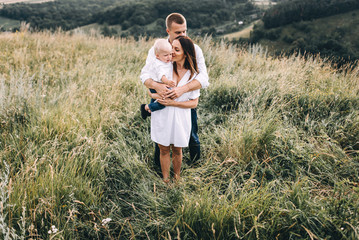 Walk beautiful young family in white clothes with a young son blond in mountainous areas with tall grass at sunset. Mother keeps son in his arms, hugging. family - this is happiness