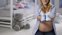Midsection Of Young Pregnant Woman With Big Naked Belly Showing Sweet Blue Baby Booties For Newborn Boy To Camera. Smiling Happy Expecting Mother Offering Baby Shoes.