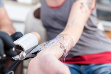 Wall Mural - Close-up of the hands of a skilled tattoo artist wearing sterile black gloves while wrapping the forearm of a tattooed client