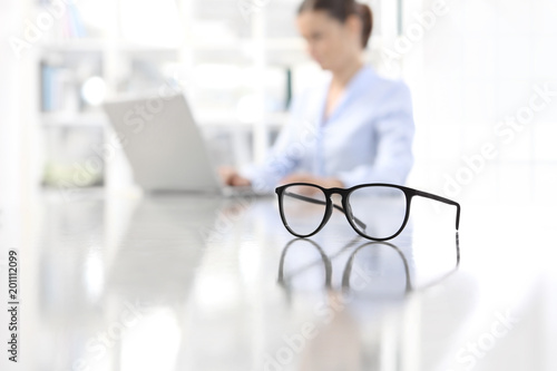 Cuadros en Lienzo eyeglasses leaning on desk and woman working on computer at office in background