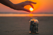 A Glass Jar With Coins And Bills On The Beach Against The Sunset. The Concept Of Accumulation Of Money For Travel