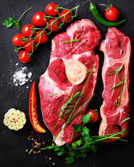 Wall Mural - Raw meat steaks with cherry tomatoes, hot pepper, garlic, oil and herbs on dark stone, concrete background. Free space for your text.