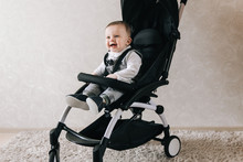 Little Smiled Boy Sits In The Baby Buggy On The Light Background