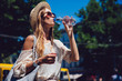 Profile of lovely woman in sunglasses, drinking a water, during walking outdoors. Dressed in trendy clothes, in hat. Summertime.