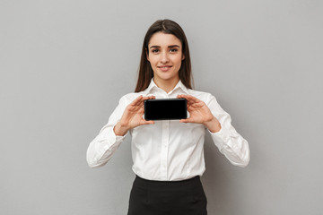 Wall Mural - Happy woman with long brown hair doing commercial and demonstrating black copyspace screen of cell phone at camera, isolated over gray background