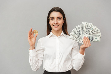 Wall Mural - Portrait of smiling successful woman in white shirt and black skirt holding bitcoin and lots of money dollar bills, isolated over gray background