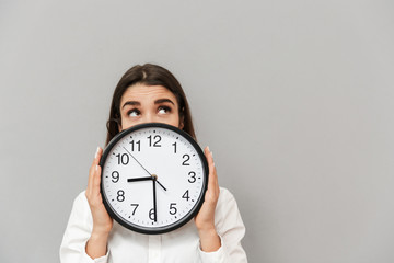 Wall Mural - Photo of business woman in white shirt looking upward while covering face with big round clock, isolated over gray background