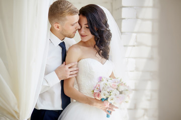 Wedding couple indoors is hugging each other. Beautiful model girl in white dress. Man in suit. Beauty bride with groom. Female and male portrait. Woman with lace veil.