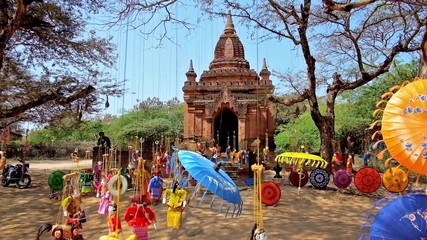 Canvas Print - BAGAN, MYANMAR - FEBRUARY 24, 2018: Traditional colored umbrellas and beautiful dolls are hanging on the tree next to the ancient Buddhist Shrine in archaeological site, on February 24 in Bagan. 