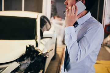 Stressful and upset young man in office business suit giving a call and talking on the phone for wrecked car crash.