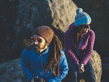 Young Couple Of Climbers Wearing Hats And Sunglasses Sharing Sunset Seated At Rocky Scenery