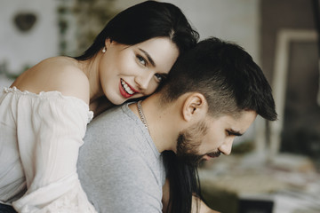 Portrait of a cute couple sitting on a chair while girl with red lips smiling is embracing his serious boyfriend.