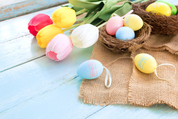 Colorful Easter eggs in nest with tulip flowers on blue wooden background.  Easter holiday in spring season, top view with copy space.