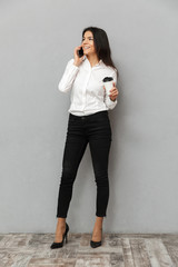 Sticker - Full length image of attractive business woman in formal wear standing and having mobile call with paper cup of takeaway coffee in hand, isolated over gray background