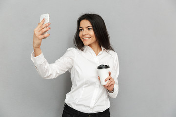 Wall Mural - Photo of secretary woman in formal wear standing holding takeaway coffee in hand and taking selfie on mobile phone, isolated over gray background