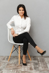 Full length image of happy business woman in formal wear sitting on chair in office, isolated over gray background