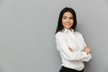 Sticker - Portrait of smiling successful woman with long brown hair wearing office clothing looking aside and standing with arms crossed, isolated over gray background