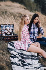 Two beautiful girls in colored shirts sitting on the grass on the hillside. Watch the sunset, drinking wine, hug, happy. picnic