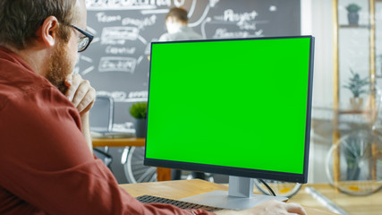 Man Works on the Personal Computer with Green Mock-up Screen. In the background Creative Office with Man Drawing on Chalkboard Wall.