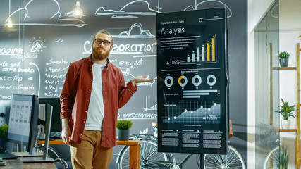 Wall Mural - Male Developer Looks at Interactive Whiteboard with Charts, Graphs and Growth Statistics. He Points at Display with Presenting Gesture and Smile. In the Background Creative Office with Chalkboard Wall