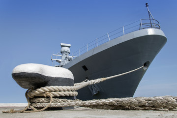 A gray modern warship moored by ropes on the shore.