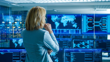 In the System Control Room Female Head of the Department Crosses Arms and Thinks Standing with Her Back to the Camera. In the Background Multiple Screens Showing Interactive Data.