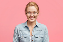 Portrait Of Pleased Attractive Smiling Female Dressed In Fashionable Denim Jacket, Sees Something Appealing In Shop, Isolated Over Pink Studio Background. Cheerful Blonde Young European Woman