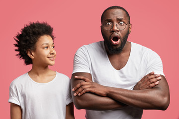 Wall Mural - Happy teenage boy with Afro hairtyle looks with positive expression at his dark skinned surprised father who keeps mouth widely opened and arms folded, shocked to recieve bad unexpected news