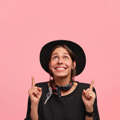 Wall Mural - Smiling beautiful young female has cowboy style, being glad after horse racing competition, looks upwards with happy expression, isolated over pink background with blank space for your hearder