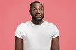 © WHstudio Leushin N - Portrait of surprised African American male in bewilderment, bites lower lip, feels puzzled, wears casual t shirt and spectacles, isolated over pink background. Dark skinned young man poses indoor
