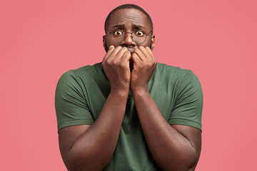 Wall Mural - Candid shot of handsome African American male looks with worried expression, bites nails, feels anxious and nervous as being scared of something or sees phobia, poses against pink background