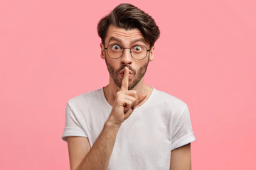 Portrait of attractive male with stubble makes silence gesture, asks not tell his secret to anybody, has eyes popped out while gossips with friend, finds out surprised recent news. Hush concept