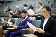 © pressmaster - Young serious businesswoman and other people in formalwear reading papers in auditorium or conference hall