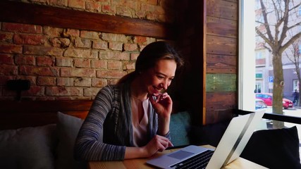 Wall Mural - Cheerful girl chatting with friends by laptop at restaurant and sitting on sofa with pillows. Concept of enjoying with social networks and wireless Internet. Young woman dressed in pink blouse and