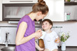 © sementsova321 - Photo of young woman in casual clothing stands against kitchen interior, looks at her adorable son with great love, gives tasty drink in mug. Positive child look at his mum with great pleasure