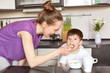 © sementsova321 - Horizontal portrait of young mother in casual outfit, feeds child with spoon, gives him healthy food, take care of son, pose at kitchen domestic interior. Motherhood, children, nutrition concept