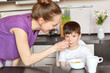 © sementsova321 - Children, family and eating concept. Portrait of delighted young cute female feeds her handsome little kid with delicious cereals or porridge, sit against kitchen interior. Bringing up child